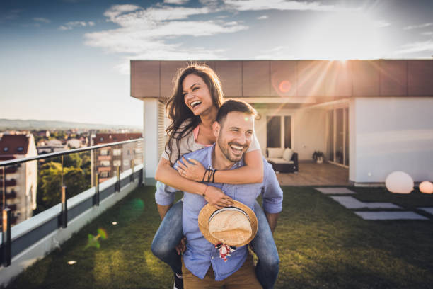 Joyful couple having fun while piggybacking on a penthouse terrace. Young playful couple having fun while piggybacking on a penthouse balcony during sunny day. apartment balcony garden stock pictures, royalty-free photos & images