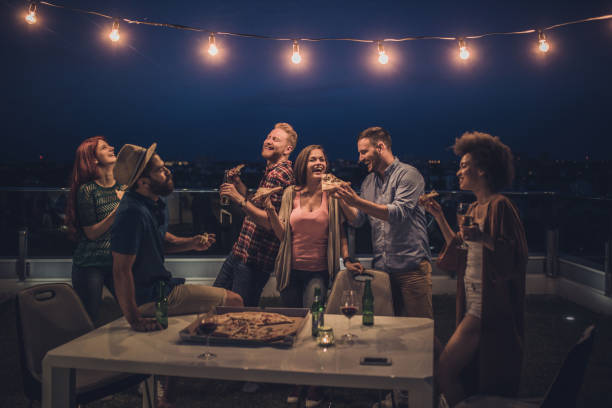 Joyful friends having a dinner party during the night on a terrace. Large group of happy friends having fun while eating pizza and drinking alcohol during night party on a penthouse terrace. apartment balcony garden stock pictures, royalty-free photos & images
