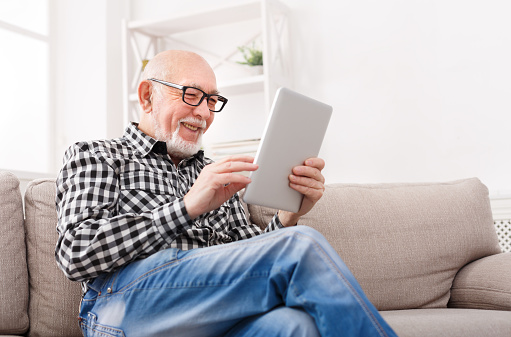 Smiling senior man reading news on digital tablet. Cheerful excited mature male using portable computer at home, copy space