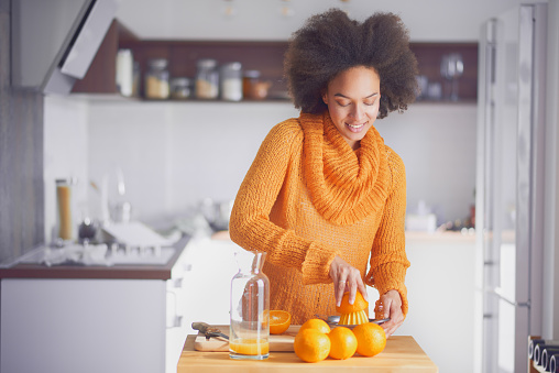 Mixed race woman squeezing orange fruit and making the juice while standing in modern kitchen