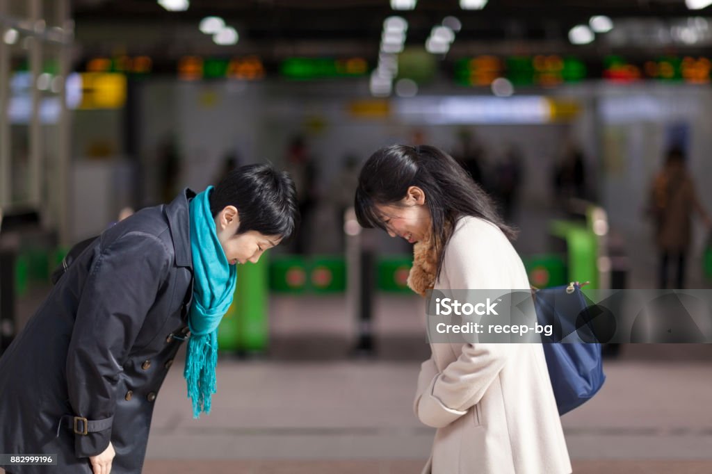 Two Japanese Businesswomen Bowing To Each Other At Station Two businesswomen are bowing to each other in front of station gates. Bowing Stock Photo