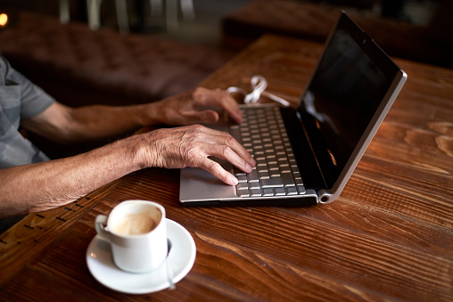Unrecognizable senior male sitting in coffee shop and having a cup of coffee while typing on laptop computer.