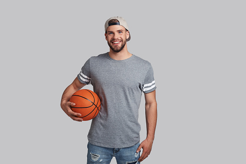 Handsome young smiling man carrying a basketball ball and looking at camera while standing against grey background