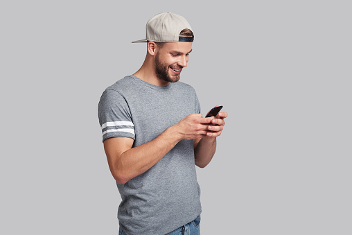 Handsome young man holding smart phone and looking at it while standing against grey background