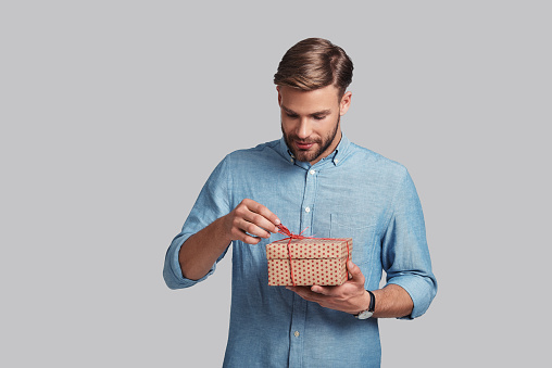 Handsome young man tying gift box and smiling while standing against grey background