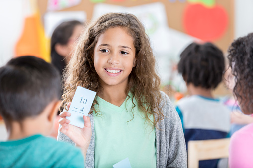 A smiling elementary age little girl stands in her after school care classroom and holds up a math subtraction flash card.  She quizzes an unrecognizable little boy.  Another unrecognizable friend looks on.