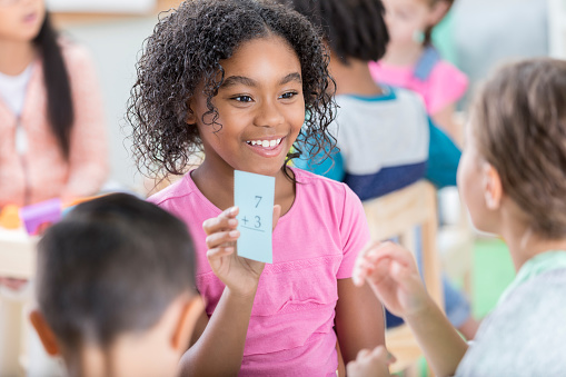 A smiling elementary age little girl sits in her after school care classroom and holds up a math addition flash card.  She quizzes an unrecognizable female friend.  They are sitting with an unrecognizable little boy.
