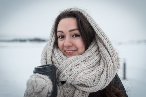 Young woman in warm and soft scarf with hot coffee in cup for take away staying at clear field on the winter background. Cold day. White and soft colours.