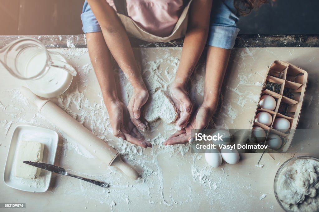 Mom with daughter on kitchen. Cropped image of attractive young woman and her little cute daughter are cooking on kitchen. Having fun together while making cakes and cookies. Top view of mom with daughter making dough heart. Cooking Stock Photo