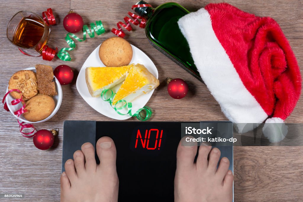 Digital scales with male feet on them and sign "no!" surrounded by Christmas decorations, sweets and bottle of alcohol. Shows how unhealthy lifestile during xmas holidays effects our body. Digital scales with male feet on them and sign "no!" surrounded by Christmas decorations, sweets and bottle of alcohol. Shows how unhealthy lifestile during xmas holidays effects our body. Top view. Over Eating Stock Photo