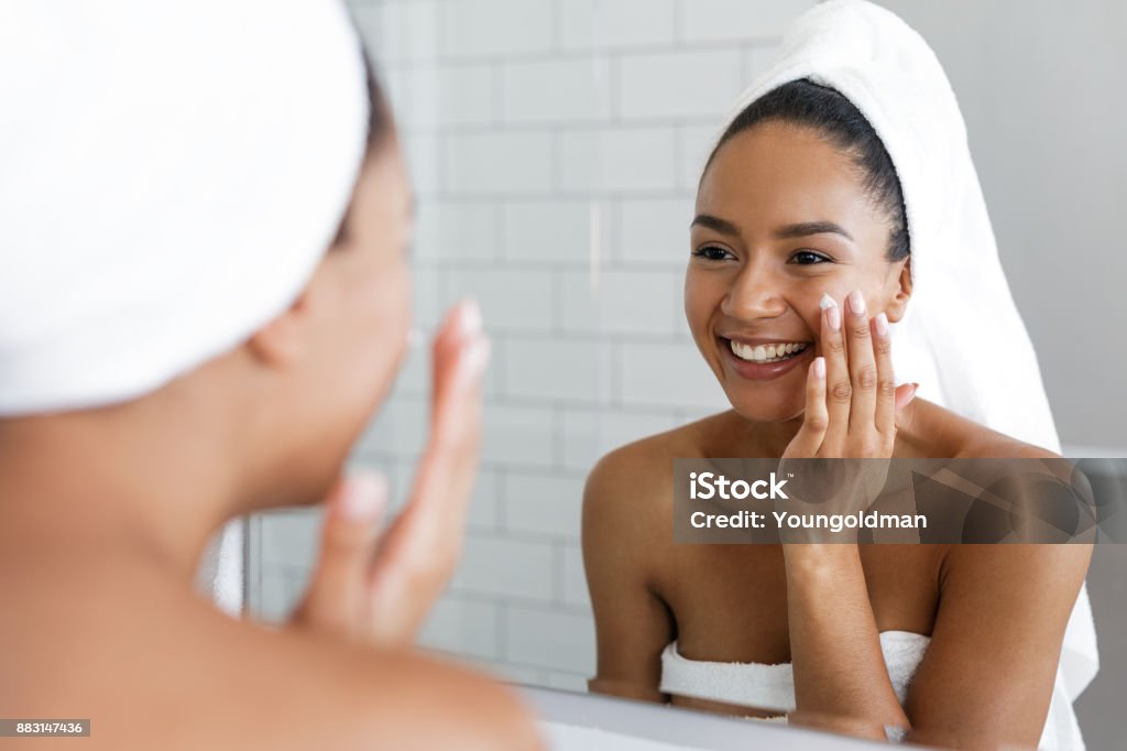 Heureuse femme mettant sur hydratant pour le visage avec la main dans la salle de bain miroir - Photo de Soin de la peau libre de droits