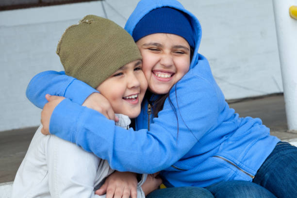 In this closeup, a teenage brother and sister sit on a set of stairs in front of a building.  The girl embraces her younger brother cheek to cheek as they both smile in the distance.