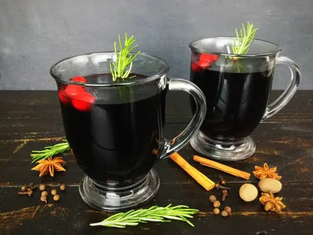 Two glass mugs filled with mulled red wine, garnished with cranberries and rosemary, and shown with various spices