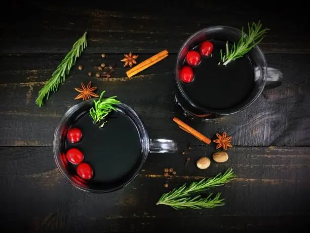 Two glass mugs filled with mulled red wine, garnished with cranberries and rosemary, and shown with various spices