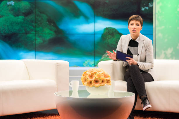 Talk show host Female talk show host sitting on a sofa, talking and gesturing, digital display in background. television host stock pictures, royalty-free photos & images