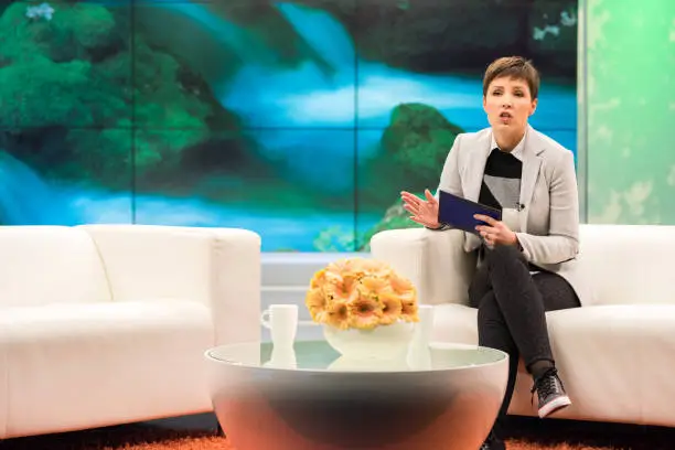 Female talk show host sitting on a sofa, talking and gesturing, digital display in background.