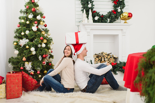 Happy cheerful funny young couple in love in red hat sitting in light room at home with decorated New Year tree and gift boxes on the background of Christmas wreath. Family, holiday 2018 concept