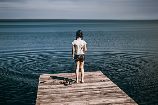 A little girl with braids stands on the bridge of the lake looking into the distance. Back view. Psychology, expectation, loneliness, return, devotion. Copy space