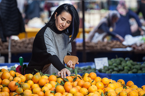 Adult woman wearing a gray sweater and sunglasses shopping in farmer's market. She is choosing oranges. She has black hair. Selective focus on model. Shot in daylight with a full frame DSLR camera.