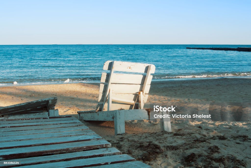One lonely old white vintage wooden sun lounger standing at the abandoned beach after the end of a summen season in the evening sunlight. Abandoned Stock Photo
