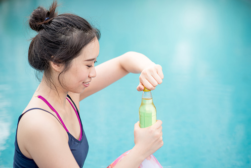 Beautiful young Asian happy woman opening sport drink water bottle after do yoga at swimming pool, outdoor exercise and healthy lifestyle concepts