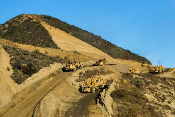 Bulldozer and excavators go through a mountain during the expansion of a road. The contrast between the blue and yellow tones of the sky and the mountains is striking.