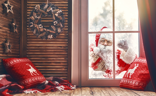 Merry Christmas! Santa Claus is knocking at window. Room decorated for holidays. View indoors home.