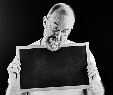 An unhappy looking mature, bearded man frowns down at a blank blackboard he's holding, full of regrets, which you can add on the chalkboard. Black and white image.