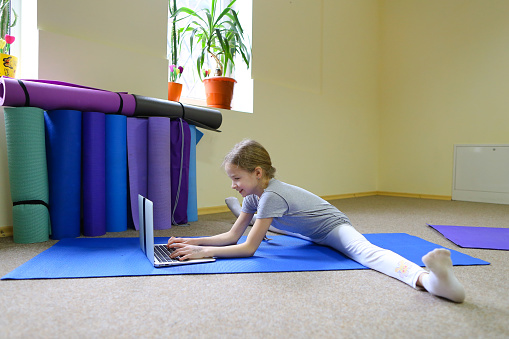 Blonde schoolgirl of European appearance sits on floor in cross-twist position and rewrites with friends through Internet on note. Child focuses on monitor and prints message. Concept of important for children to have healthy lifestyle from early years, or in ability to communicate with loved ones through Internet.