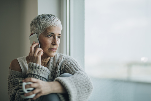 Thoughtful mature woman talking to someone over smart phone and looking through window. Copy space.