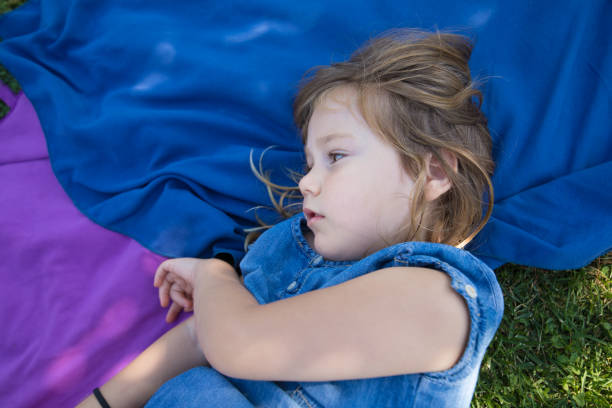 four years old blonde girl lying on towels in the green grass of park with thoughtful or imagining face