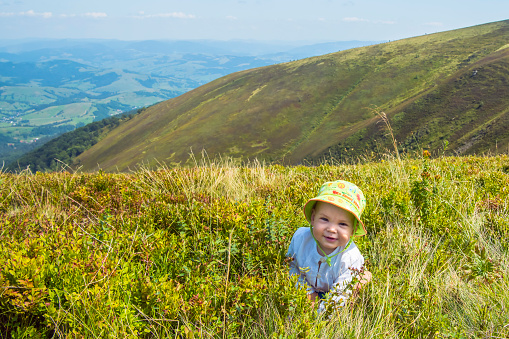 Smiling baby playing with grass in the mountains in summer. Beautiful mountains landscape in the background.