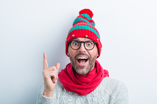 Funny christmas portrait of nerdy man wearing elf cap and red scarf. Excited man wearing winter sweater pointing with index finger against white background.