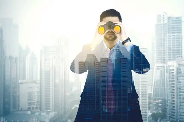 Double exposure of young businessman looking through binoculars with modern city background