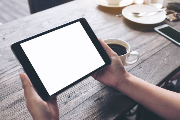 immagine mockup delle mani di una donna d'affari che tiene il tablet pc nero con schermo bianco bianco bianco e tazza di caffè su tavolo di legno vintage sullo sfondo del caffè - pc desktop pc women desk foto e immagini stock