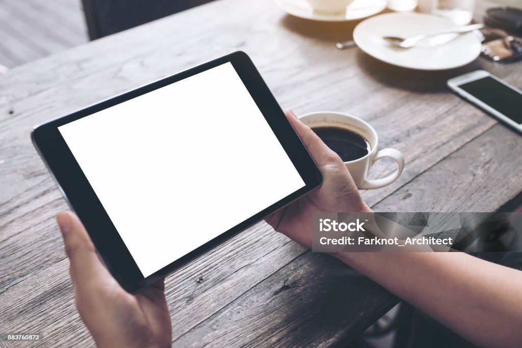 カフェ背景で黒いタブレット pc、空白の画面とヴィンテージの木製テーブルの上のコーヒー カップを保持しているビジネスの女性の手の実物大模型のイメージ - タブレット端末のロイヤリティフリーストックフォト