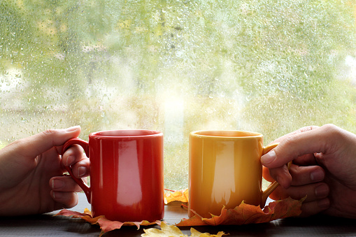 pair of hands holds hot drink in the colored mugs on the background window with raindrops