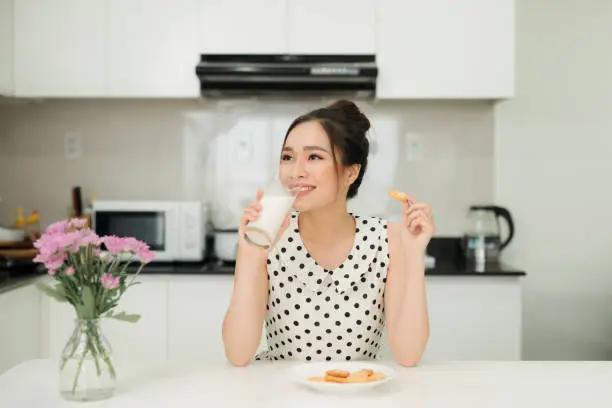 Photo of Young asian woman holding milk glass bite cookie in her kitchen