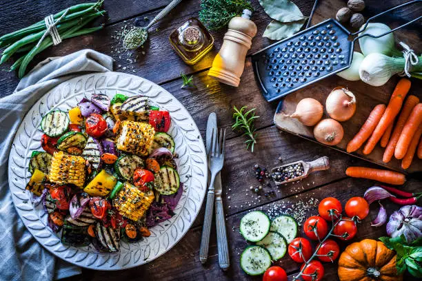 Grilled vegetables plate shot from above on rustic wooden kitchen table. The plate is on a gray textile napkin at the left of an horizontal frame and some fresh vegetables are at the right. DSRL studio photo taken with Canon EOS 5D Mk II and Canon EF 100mm f/2.8L Macro IS USM