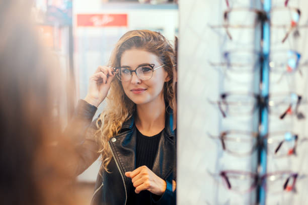 sorridente giovane donna che prova gli occhiali sullo specchio in ottica. - misurare foto e immagini stock