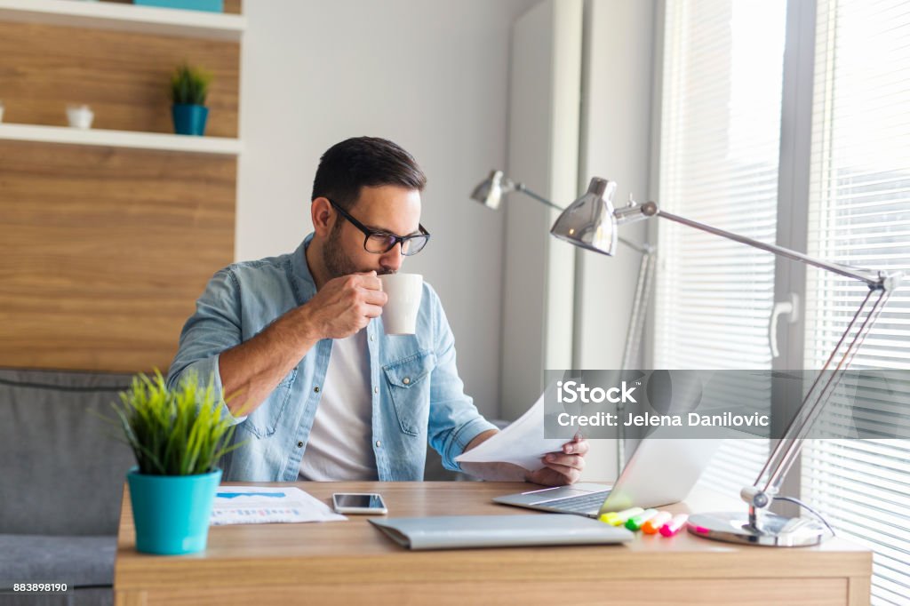 Remote job Young relaxed freelancer drinking coffee and looking at some documents Adult Stock Photo