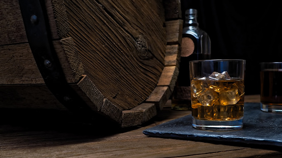 Two glasses and bottle of whiskey on wooden bar counter near oak whiskey barrel. Black background