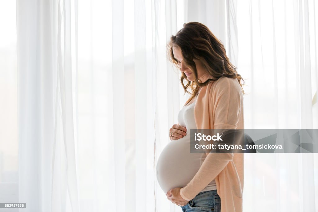 Portrait of young pregnant attractive woman, standing by the window Portrait of young pregnant attractive woman, standing by the window, dressed in casual clothing, day before due date Pregnant Stock Photo