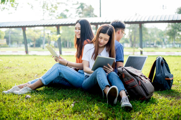 Group of Asian college student using tablet and laptop on grass field at outdoors. Technology and Education learning concept. Future Technology and Modern entertainment concept. Edutainment theme.