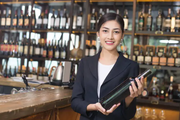 Young Asian Woman Restaurant Catering Service  with Happy Emotion. Woman Present Red-Wine for Customer at Bar. Woman with Wine in Bar Concept.