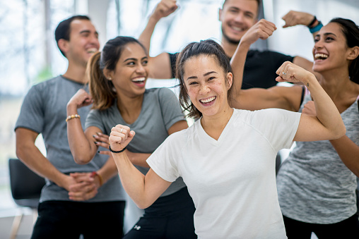 A multi-ethnic group of adult men and women are indoors in a health center. They are laughing together while flexing their muscles.