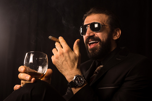 The laughing successful elegant businessman in black suit is smoking cigar and drinking alcohol on black background.