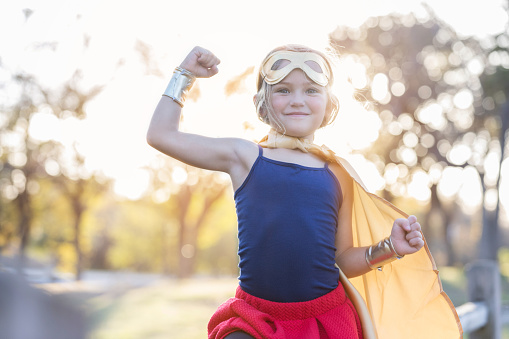 Little girl enjoys dressing up as a superhero. She is smiling at the camera while flexing her muscles.