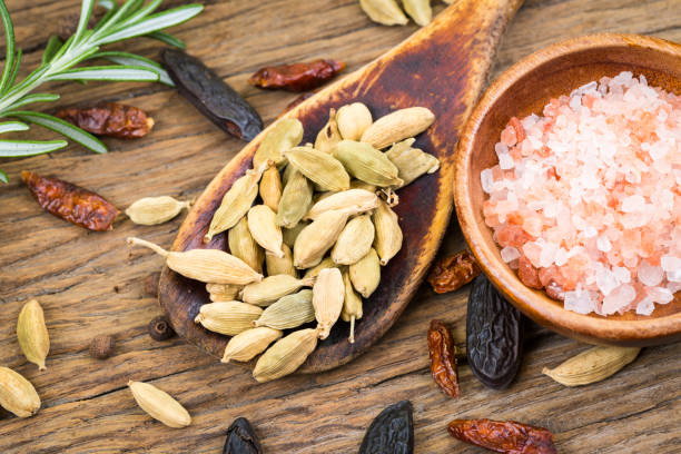 closeup of whole cardamom on a wooden spoon and pink crystal salt in a small wooden bowl on rustic wood background and a sprig of rosemary - salt crystal spoon food imagens e fotografias de stock