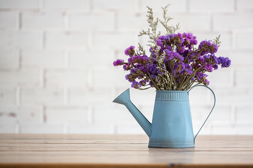 Violet dried flowers in blue tin watering flower pot on wooden table with white brick wall background.